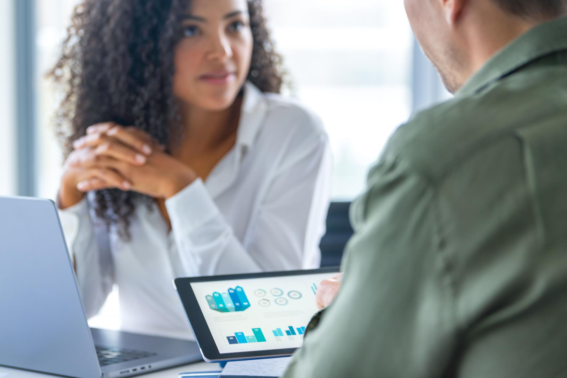 Business man and business woman in a meeting at the office. There is a laptop on the table and the man is holding a digital tablet with finance chart and graphs.